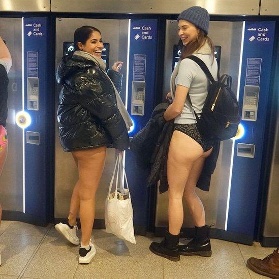 Two women smile as they use station ticket machines wearing coats and pants.