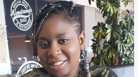 Edwige Nsilu sitting in front of a house plant at a bar. She has black hair, with braids that have white highlights in them. She is wearing a necklace and a green camoflague top and is smiling at the camera.