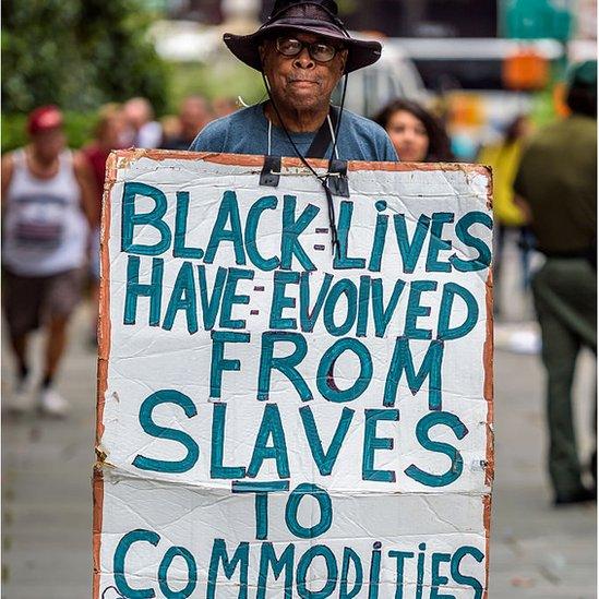 Activist holding sign reading BLACK LIVES HAVE EVOLVED FROM SLAVES TO COMMODITIES