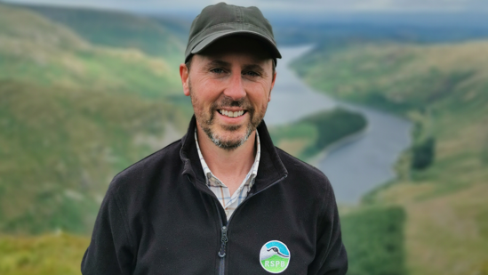 Mr Swainson is wearing an "RSPB" branded black fleece and a dark green cap. He has a short brown/ grey beard and is standing in front of a rolling green landscape with a reservoir in the background.