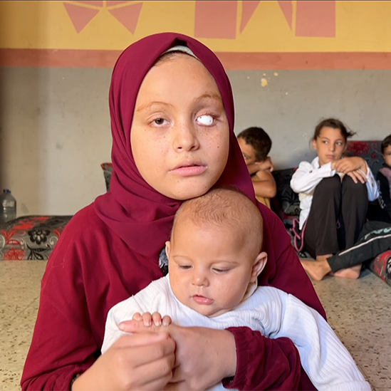 A young teenage girl - Malak - holds her baby sister, Rahma, on her lap. Malak is wearing a burgundy headscarf and long-sleeve tunic. One of her eyes has been replaced with a white sphere. She is looking to the side of the camera. The baby is holding onto her sister's finger and looking at it. She is wearing a long-sleeved white top. In the background, their five siblings sit close together in the corner of a bare room with its walls painted with square patterns,  on large floor cushions. 