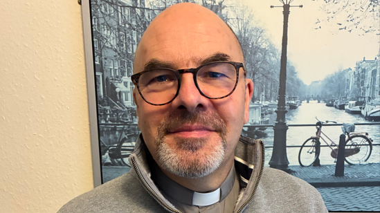 Richard has very closely cropped hair and a greying beard and glasses. He is wearing his church clothing with a beige jumper on top.