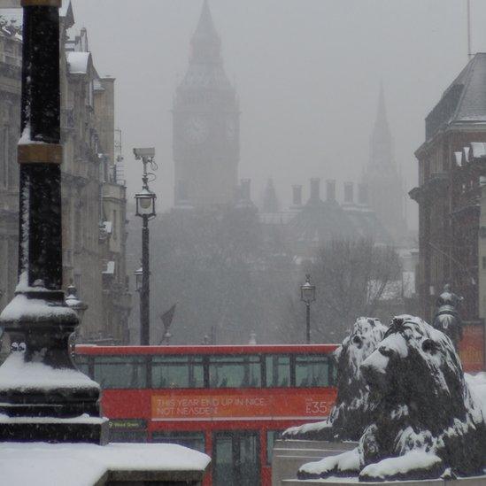 In the foreground are two lion statues, covered in snow and behind is a red London bus. In the background, Big Ben and the Houses of Parliament can just about be seen in the fog.