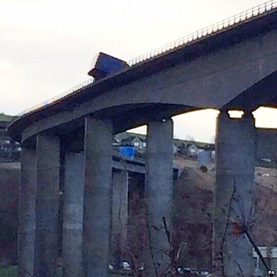 A lorry on its side on a bridge, part of it hanging over the edge