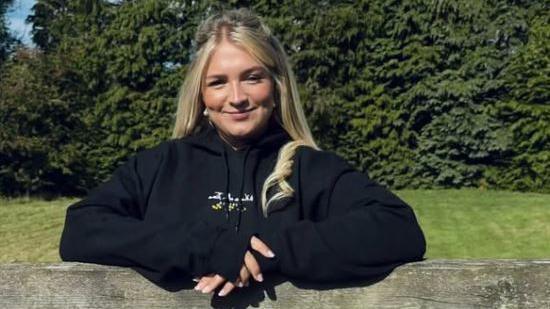 Young blonde haired woman in a black hooded top. She is leaning on a gate and smiling. There is a green grass lawn and fir trees behind her and it is a bright day. 
