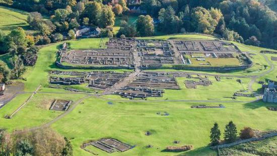 An aerial view of Vindolanda with the Roman remains of Roman walls visible and surrounded by green fields.