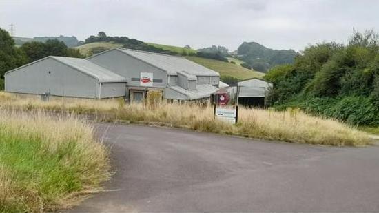 An industrial building in a rural setting. The large grey building is surrounded by dry grass and there is a road leading to it. 