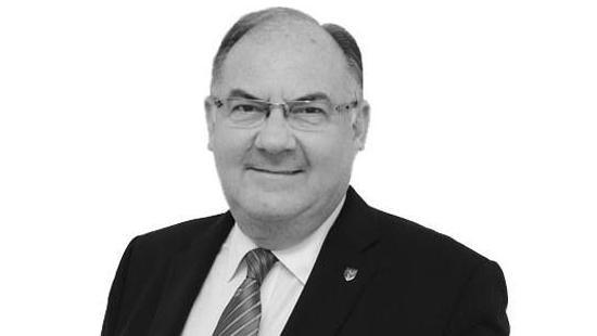 A black and white image of Geoff Corkish smiling. He has dark hair with a receding hairline and glasses and is wearing a dark suit jacket over a white shirt with a striped tie. There is a shield shaped pin badge with the Three-Legs-of-Man symbol on it on his lapel.