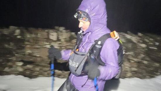 A woman wearing a purple hooded coat and rucksack treks across a snowy terrain with walking poles. A dry stone wall is in the background and it is dark.