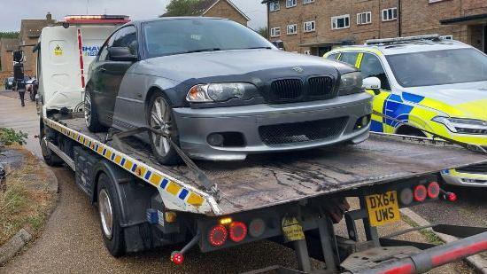 A BMW on the back of a removal truck. It is a dark grey car with a light grey panel across the bonnet. To the side of the truck is a police car.