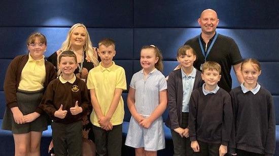 Seven children from two schools stand with their teachers at the opera event. One pupil, wearing a brown jumper and yellow T-shirt, is giving a thumbs up to the camera. Three pupils have a yellow and brown uniform with a teacher who has long blonde hair, four are in blue and standing with their teacher, who has a blue lanyard around his neck and has a bald head.