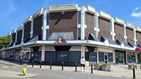 A brick building with slit windows and glass canopies above large shop windows, many of which have a grey covering over them