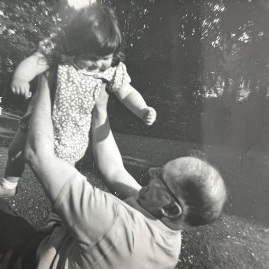 A black and white photo of Anthony Pratt holding his daughter in the air as a young girl. He is lying on the grass, with trees in the background