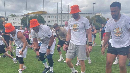 People doing burpees on a rugby pitch. Four men are in the foreground in Leo and Friends T-shirts, and three of them are also wearing orange bucket hats. Behind them, several other participants and spectators are visible, with a long white three-storey building behind them.