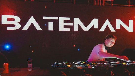 A man in a white T-shirt smiling whilst DJing. There is a dark background but large letters saying 'Bateman' above him in white.