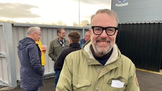 Shane Whaley, wearing a khaki-coloured coat, black jumper, and black-rimmed glasses, stands in front of a black metal shed and in front of a small group of fellow fans.