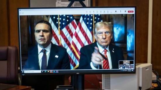 Donald Trump and his lawyer Todd Blanche (left) appear virtually on a computer desktop screen from Florida for his sentencing in his Manhattan hush money case. Trump has his finger pointing at the camera, with US flags positioned behind him and Blanche.