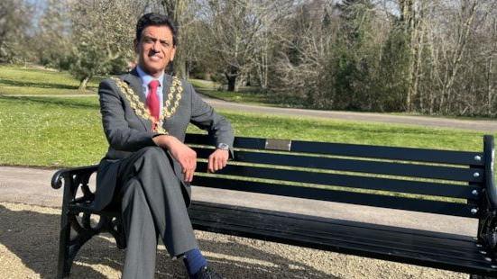 The Mayor of Oldham Dr Zahid Chauhan sits on a bench wearing his mayoral chain. A green and the trees of Alexandra Park can be seen in the view behind him. 