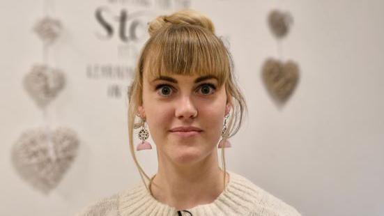 A young woman with blonde, with a full fringe and her hair tied up, wearing decorative earrings and a cream jumper.