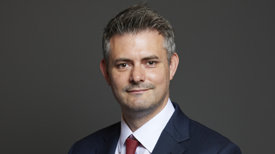 Man with short grey hair, blue suit jacket and red tie stood against grey background