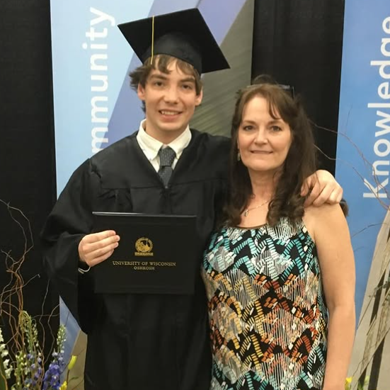 Jared puts his arm around his mother's shoulders and holds a plaque in the other. He wears a graduation cap and gown and has short dark hair. Kelly has long brown hair and wears a multi-coloured patterned dress with blue and orange tones. 