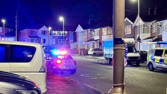 A police car with its blue lights on in a residential street at night. There is police tape attached to a lamppost and another police vehicle parked nearby.