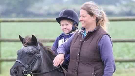 Ms Ackland stands next to a dark coloured pony that is being ridden by her child. She is patting the horses neck and looking into the distance. The child is smiling and wears a riding helmet.