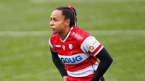 Jade Shekells stands on the pitch with her hands on her hips during a game for Gloucester-Hartpury. She wears a Gloucester-Hartpury shirt with a long sleeved black top covering her arms.