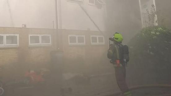 A firefighter in protective gear points a hose at a burning residential building. He is surrounded by smoke.
