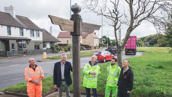 A group of men, some wearing high visibility jackets, stand around a sign post at the side of a road.