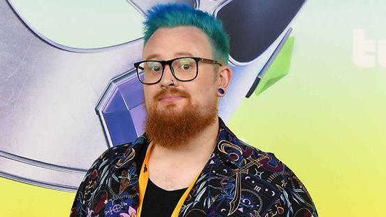 A person looks at the camera with dyed blue hair and a ginger beard. They're wearing glasses and have a nose piercing