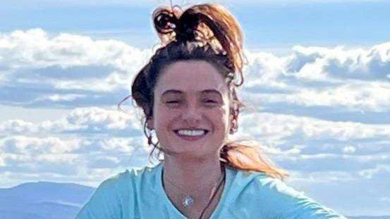 Eleanor Thompson is pictured sitting on a rock, with the landscape below, wearing a blue top and jeans, with a jumper tied round her waist. Her hair is tied in a high ponytail and she is smiling.