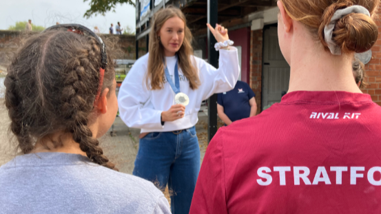 Esme speaking to youngsters at the club while holding her medal. We see the backs of two girls, one with her brown hair in braids, the other with her auburn hair in a bun, looking towards Esme.