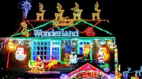 A house covered in lots of colourful festive lights, including ones of Santa, reindeer on the roof, and stars. There are also ones that say "Merry Christmas" and "wonderland".