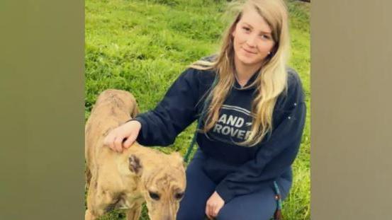 A young woman with long blonde hair is wearing a navy Land Rover hoodie and navy leggings as she sits in a field with a dog, smiling.