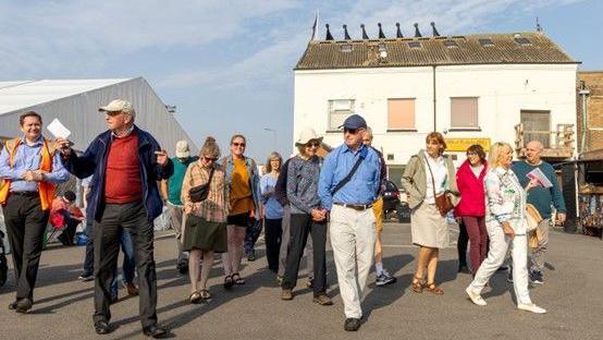 A crowd of people taking part in a guided walk. There is a white building in the background with boarded-up windows.