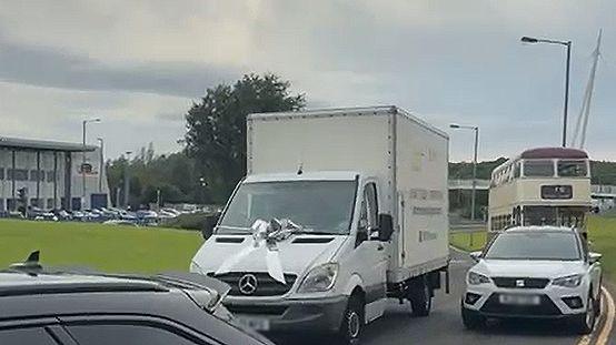 A white removals van with a silver bow on the bonnet overtakes a white car and a vintage double-decker bus.
