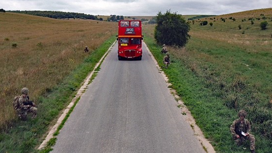 A red double-decker Routemaster bus travels along an unmarked road. Passengers can be seen sitting on the upper deck of the bus, which has an open top. The road is flanked by five soldiers wearing camouflage uniforms and kneeling in the grass verges