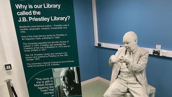A sign on the left explains why the University of Bradford's library is named for the writer J B Priestley and next to the sign is a white statue of the author seated and holding a pipe in his hands