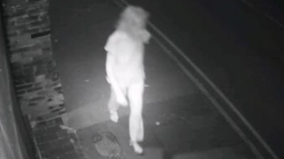 Black and white CCTV image of woman in white pyjamas walking on pavement towards the camera, with double painted lines visible on the road.