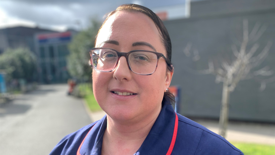 Ms Walford, in glasses, is wearing her blue uniform and is standing outside her place of work. 