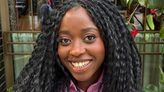 Minah Shannon has long black wavy hair and is wearing a purple shirt. She is smiling at the camera.