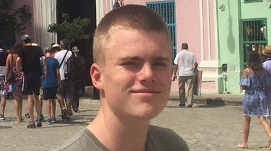 Christian Hobbs, slightly side on to the camera. He was wearing a khaki coloured t-shirt and is smiling slightly. He had cropped light brown hair. Behind him were cobbles and a mix of different people in summer clothes walking towards a street entry