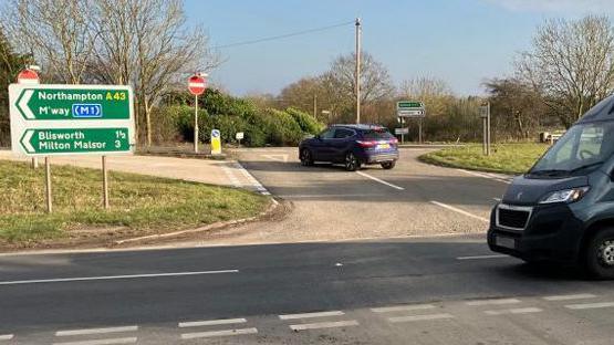 A road junction, showing a grey van, driving along the road, and a blue car at a junction. You can see several road signs and trees. 