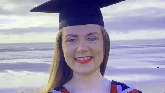 Francesca Murphy wearing graduation clothes and cap, standing, looking into camera with Swansea beach behind her. She is smiling.