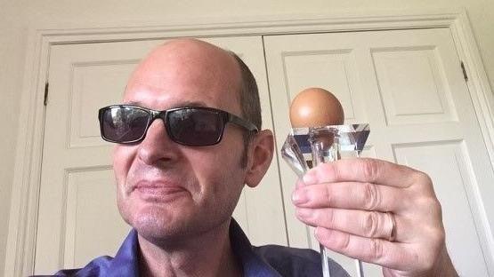 Ed  Pownall has a bald head and is wearing sunglasses with black frames. He is holding the egg on a transparent stand in his left hand, up close to his face, as he smiles towards the camera.