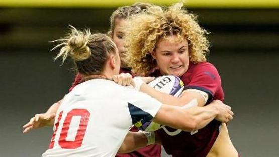 Three women fight for the ball in a game of rugby. They are standing upright and one is clutching the ball while another tries to pull it off her.