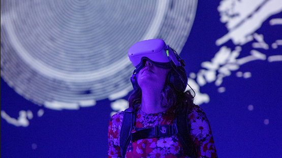 Woman wearing a VR headset at the exhibition, gazing upwards in front of a blue and white exhibition display