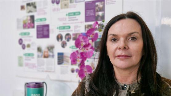 A woman with dark, long hair, smiles at the camera as she sits in an office, with a RASA charity tin beside her, and a RASA noticeboard in the background.