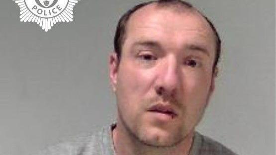 A man with black receding hair is photographed in a grey sweatshirt in front of a grey background. 